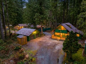 A cabin complex surrounded by dense forest, featuring multiple wooden cabins with attached garages, a central larger cabin with a porch, and a gravel driveway. - Mountain Retreat -Hot Tub -Fire Pit -Stevens Pass -River Access -Pet Friendly (Index)