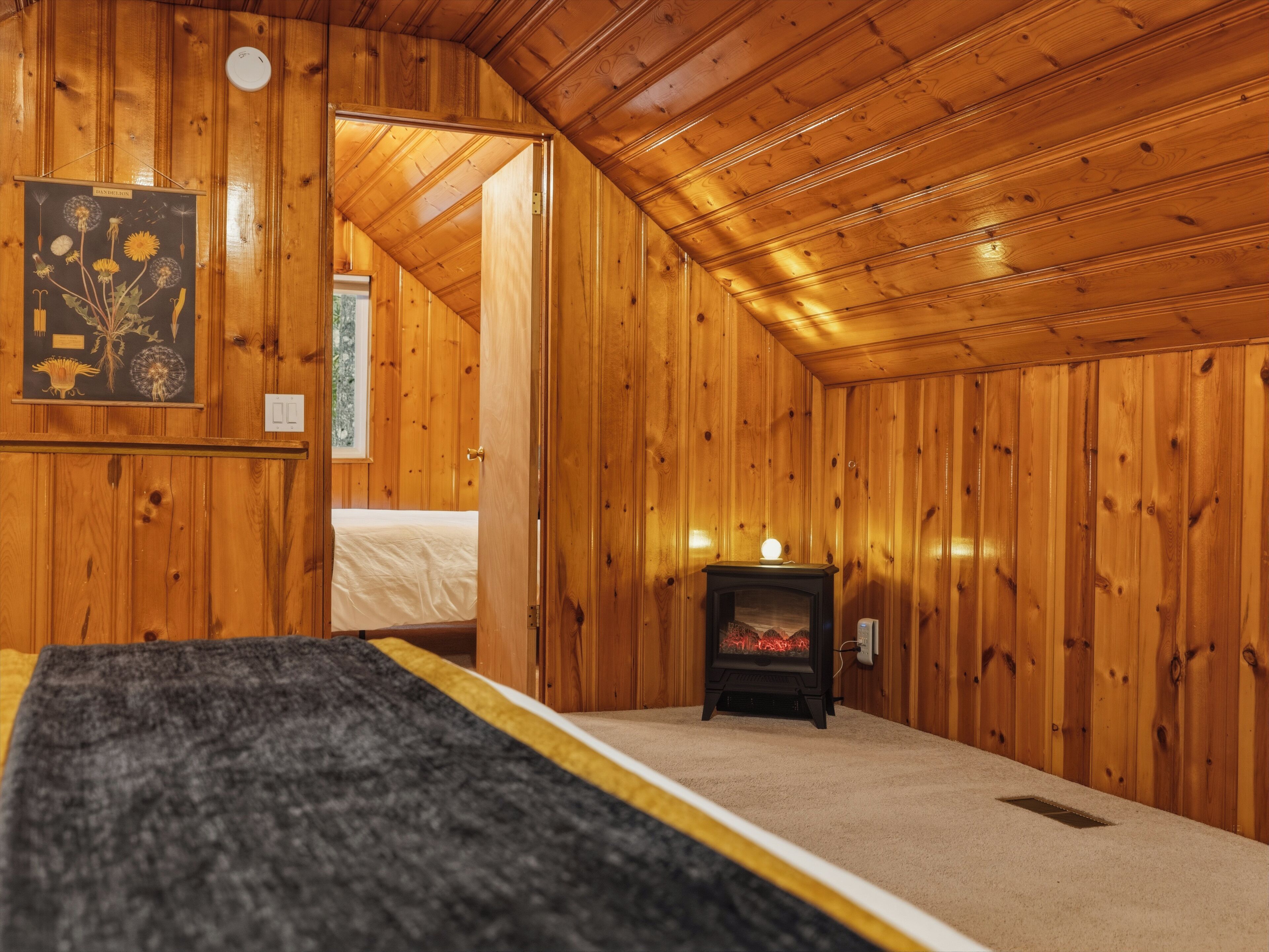 Interior de una cabaña de madera con una cama, una chimenea e ilustración botánica en la pared.