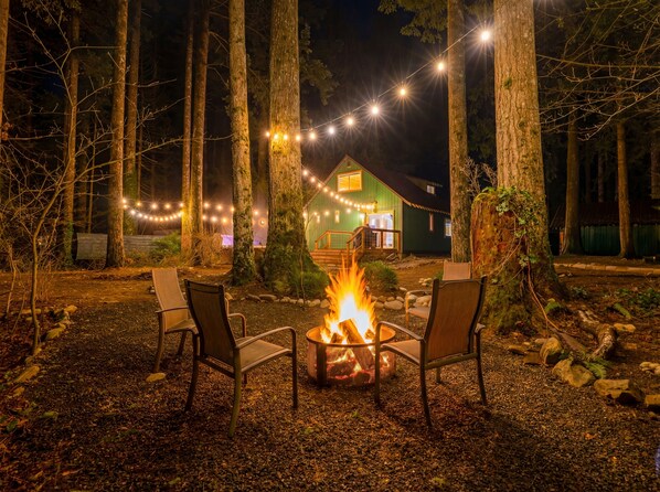 A campfire with chairs in a forested area, string lights, and a green building in the background. - Mountain Retreat -Hot Tub -Fire Pit -Stevens Pass -River Access -Pet Friendly (Index)