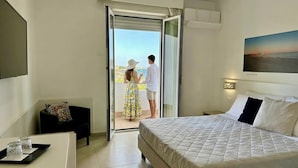 A hotel room with a bed, a desk with a tray of glasses, a sofa with a patterned cushion, and a sliding glass door leading to a balcony. - HOTEL JONIO (Noto)