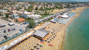 An aerial view of a coastal town with a long beach lined with rows of umbrellas and sun loungers. There are buildings and a road visible in the background. - HOTEL JONIO (Noto)