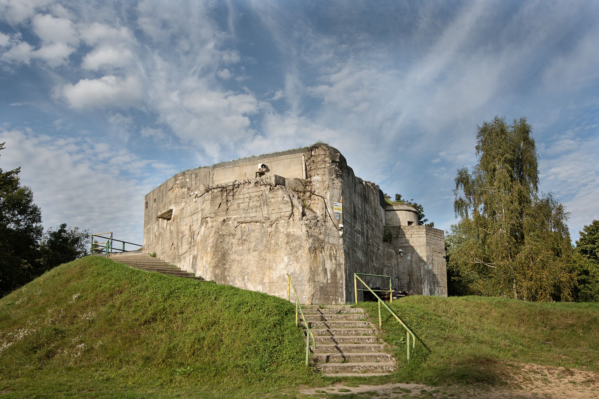 A large, historic stone building with a staircase leading up to it, surrounded by grass and trees.