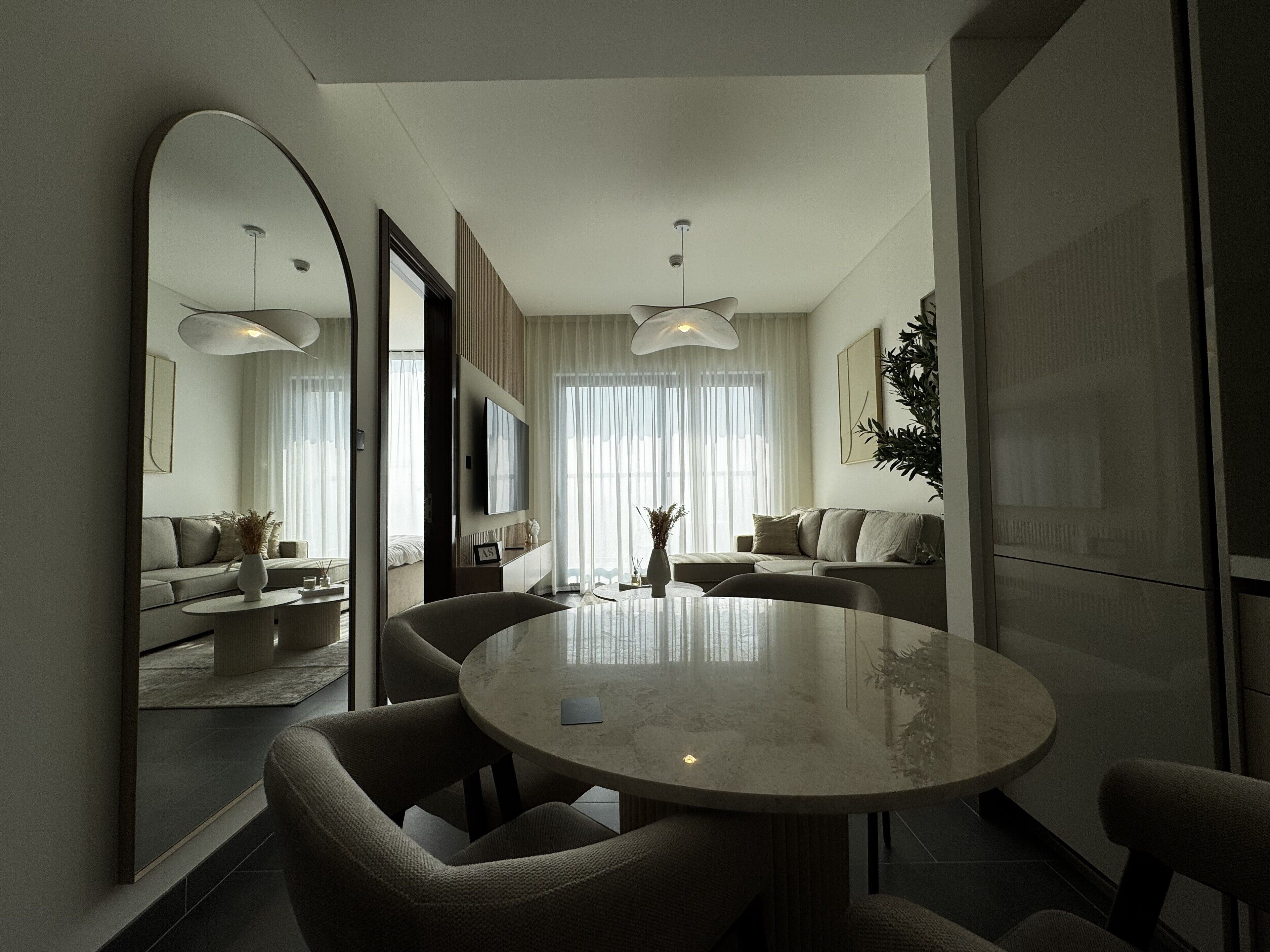 A modern living room with a round dining table, comfortable seating, a large mirror, and a view of a bedroom with a bed and curtains.