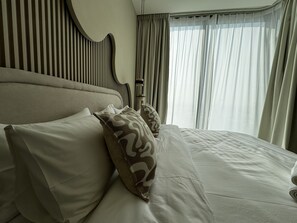 A neatly made bed with pillows, a headboard with vertical slats, and sheer curtains on a window. - Luxury 1BR Apartment with Pool Access (Dubai)