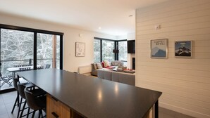 A modern kitchen with a large island, a dining area with chairs, a sofa, and a fireplace. There are large windows offering a view of trees. - Le Repère du Sommet - Spa (Orford)
