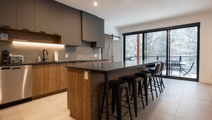 A modern kitchen with a central island, bar stools, and a view of a snowy landscape through large windows. - Le Repère du Sommet - Spa (Orford)