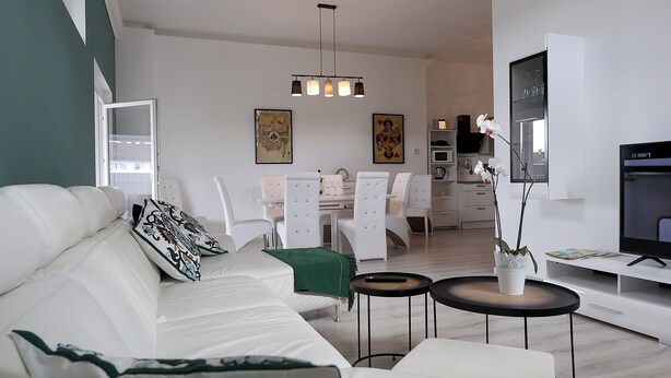 Een moderne woonkamer met een witte bank, een eethoek met witte stoelen, een keuken op de achtergrond en een tv op een dressoir.