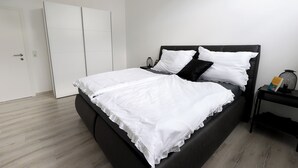Een netjes opgemaakt bed met witte beddengoed, een zwart hoofdeinde en een nachtkastje met een lamp en een boek.