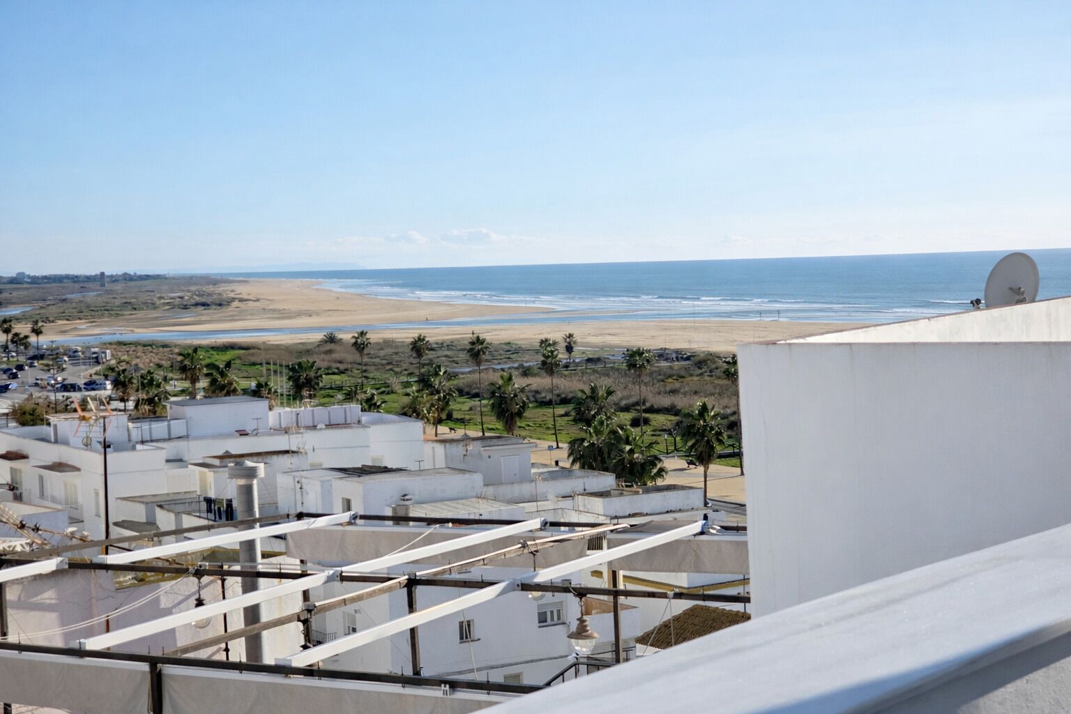 A coastal view with a beach, palm trees, and a white building with multiple water tanks and satellite dishes.
