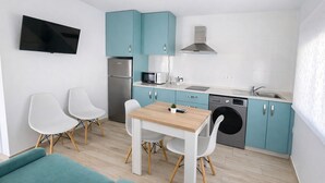 Eine kleine Küche mit türkis-blauen Schränken, einem Edelstahl-Kühlschrank, einer Mikrowelle, einem Geschirrspüler und einem Holztisch mit zwei weißen Stühlen.