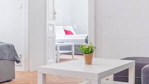 A modern, minimalist room with a white bed, a small table with a plant, and a grey sofa. - Stylish 2BR Apt Near AKH Easy Center (Vienna)