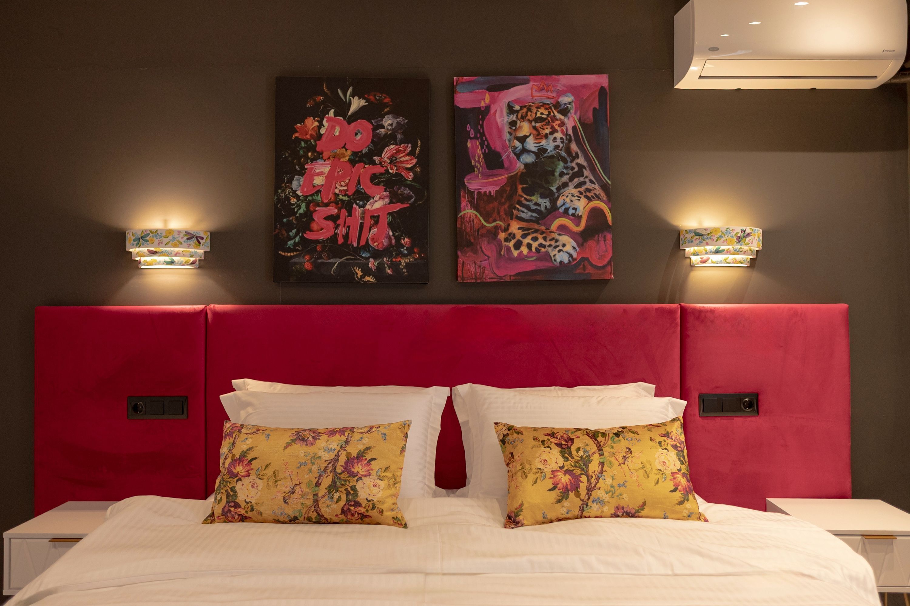 Habitación de hotel con cabecera roja, dos lámparas de pared y dos almohadas estampadas con motivos florales sobre una cama.