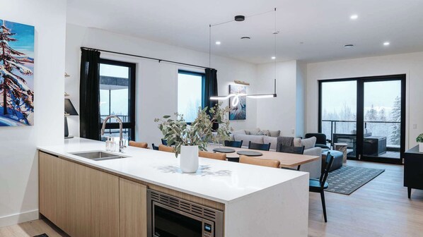 A modern kitchen with a central island, a dining area with a sofa, and large windows offering a view. - Revelstoke - Gary's Birthday- New condo! (Revelstoke)