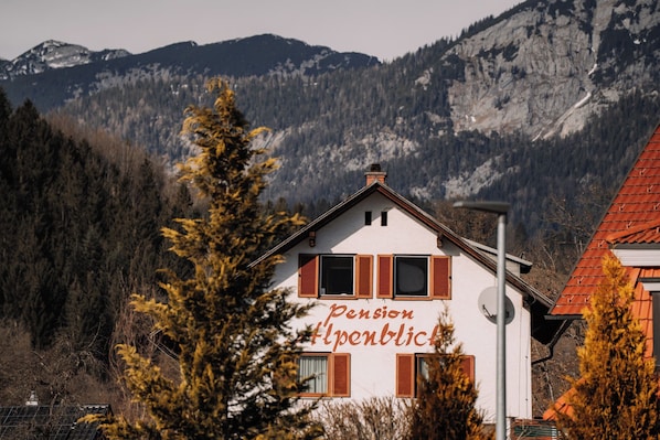 A building with the sign "Pension Alpenblick" in front of a mountainous landscape.