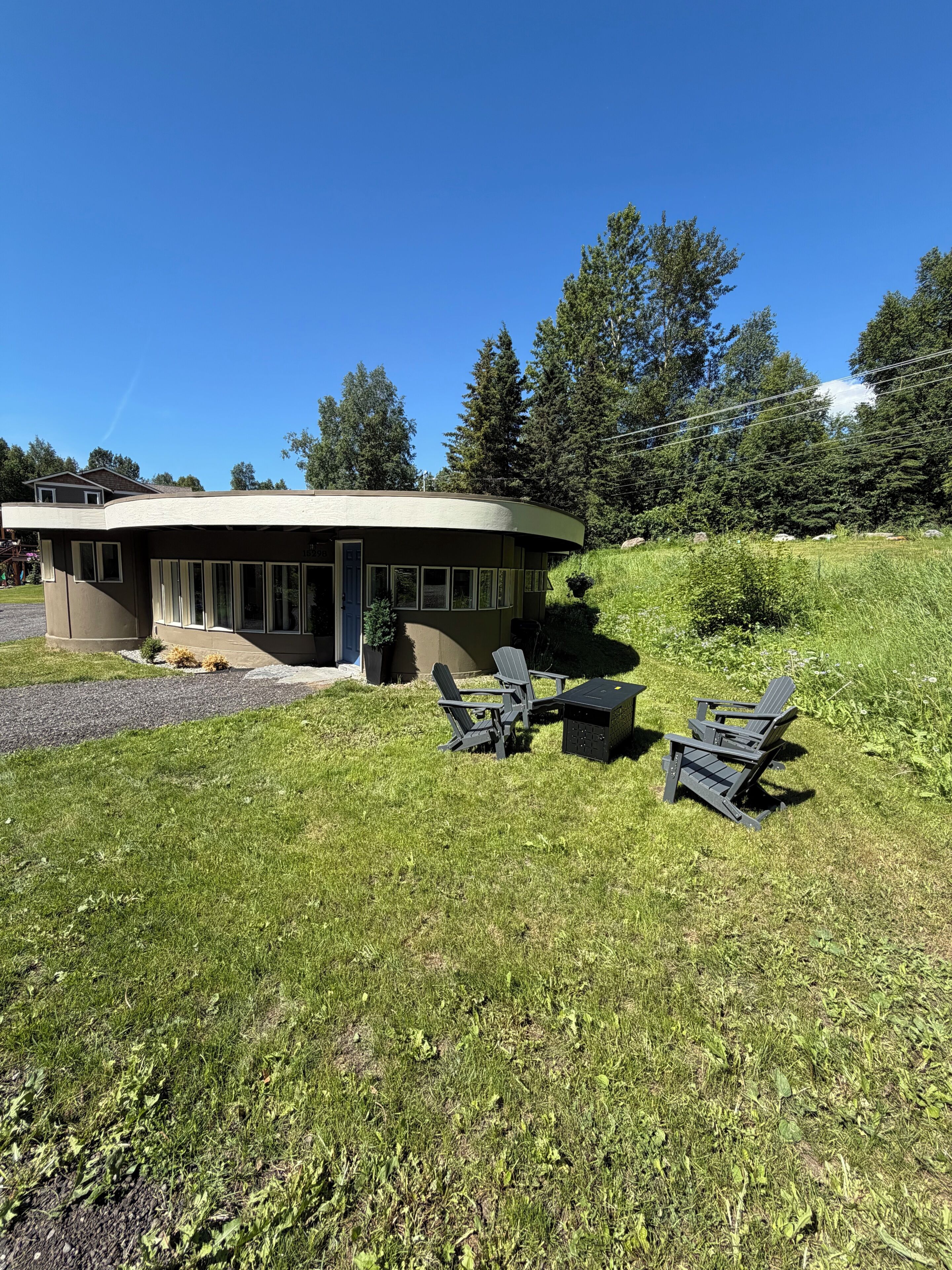 A modern building with a curved roof, a patio with Adirondack chairs, and a grassy area.