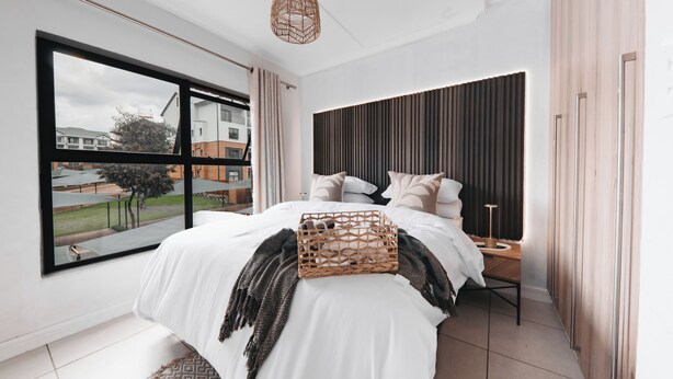 A modern bedroom with a large window, a bed with white bedding, a woven basket, and a bedside table with a lamp. - Casa De Lior- Luxury Apartment (Sandton)