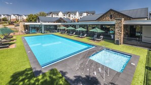 A swimming pool with a water fountain, surrounded by lounge chairs and umbrellas, with a view of residential buildings in the background. - Casa De Lior- Luxury Apartment (Sandton)