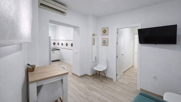 Ein modernes, helles Zimmer mit einem Holztisch, einem weißen Stuhl, einem türkisblauen Sofa, einem an der Wand befestigten Flachbildfernseher und einer Küchenzeile mit weißen Schränken und einer gefliesten Rückwand.
