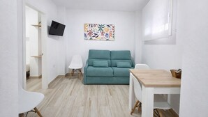Ein kleiner, heller Raum mit einem türkisblauen Sofa, einem weißen Sessel, einem Holztisch und einem an der Wand befestigten Flachbildfernseher.
