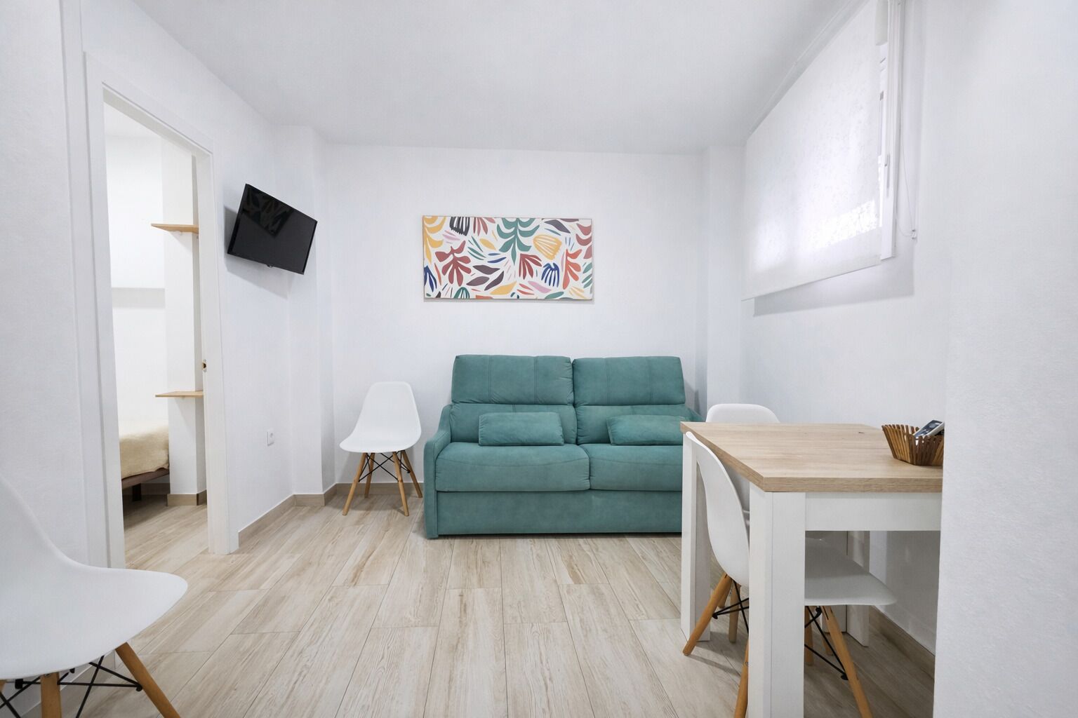 Een compacte, moderne woonkamer met een blauwgroene bank, een witte fauteuil, een houten tafel en een kleurrijk abstract schilderij aan de muur.