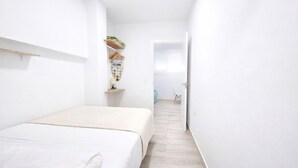 Ein schmales Zimmer mit weißen Wänden, einem Bett, einem kleinen Regal mit Kleidung und einer Tür zu einem weiteren Raum.