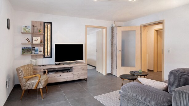 Una sala de estar moderna con televisor, mueble de entretenimiento de madera, una silla, un sofá y una mesita.