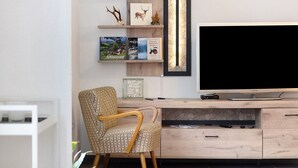 Una sala moderna con un televisor de pantalla plana grande, estantes de madera con objetos decorativos, un sillón estampado y una consola con cajones.