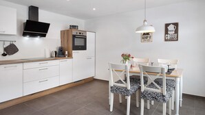 Una cocina moderna con armarios blancos, horno empotrado y televisor sobre la estufa. Hay una zona de comedor con una mesa y sillas, y dos letreros en la pared que dicen 'WHIP IT' y 'COFFEE'.