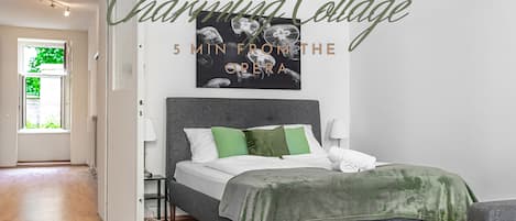 A modern bedroom with a grey bed, green pillows, and a grey headboard. There is a framed picture on the wall and a window with a view of greenery.
