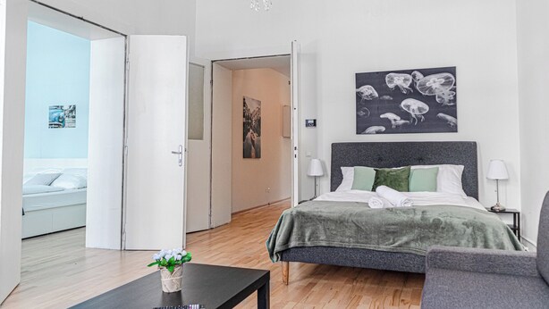 A modern bedroom with a bed, bedside tables, a chandelier, and a view of another room. - Cute 2BR Apt Near AKH Hospital Central (Vienna)