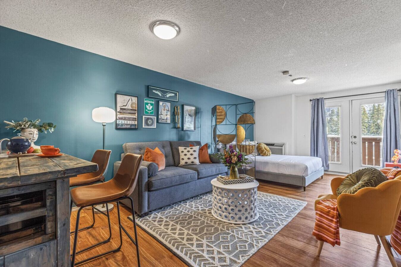 A modern living room with a blue accent wall, a sofa, a dining table with chairs, a coffee table, and a bed with a balcony view.