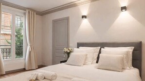 Una cama bien hecha con sábanas blancas, un jarrón con flores y una ventana con cortinas.