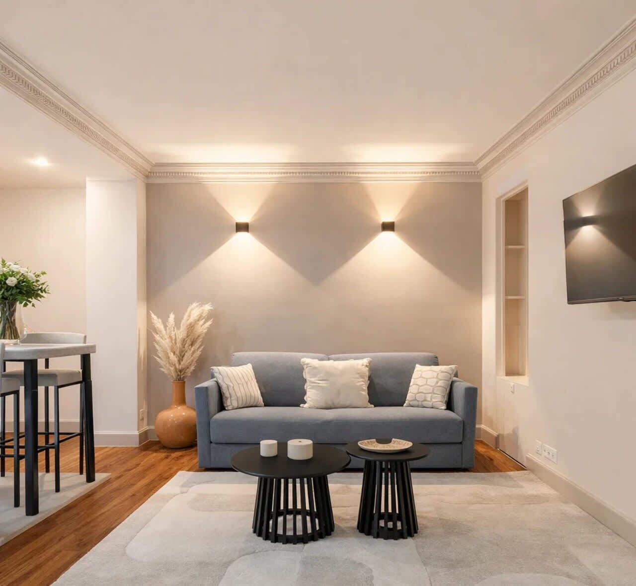 Un salon moderne avec un canapé gris, deux tables noires, un vase contenant de l’herbe des pampas et un téléviseur fixé au mur.