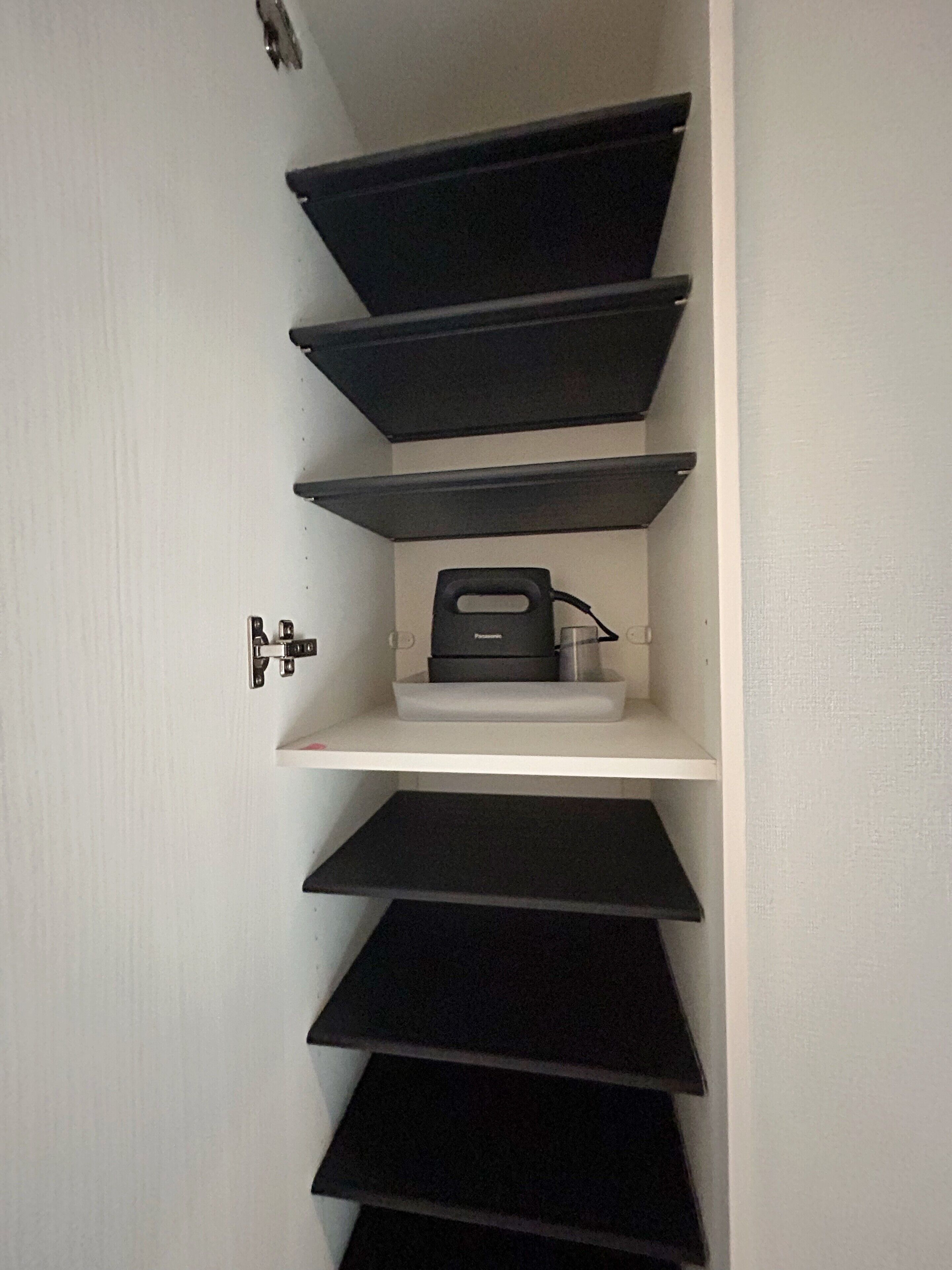 A white wall with a black shelf unit containing a black object, possibly a printer, and a small white shelf below it.