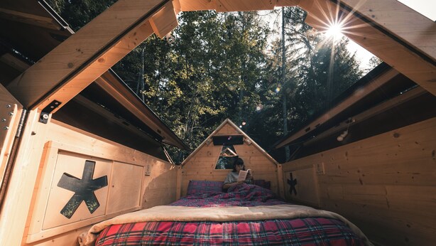 Une cabane en bois chaleureuse, dotée d’un toit en pente, d’un lit recouvert d’une couverture à carreaux et d’une grande fenêtre laissant entrer la lumière du soleil.