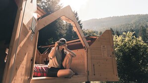 Une cabane en bois avec un toit en pente, un lit recouvert d’une couverture à carreaux et une vue sur une colline boisée.