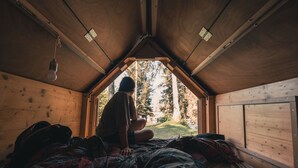 Un chalet confortable avec un plafond en pente, un lit recouvert d’une couette à carreaux et une vue sur une forêt, visible à travers une fenêtre triangulaire.