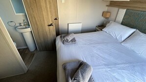 Quarto de hotel com cama, mesa de cabeceira com abajur, guarda-roupa de madeira, banheiro com pia e vaso sanitário, e radiador.
