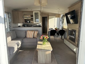 A compact living space with a sofa, a small wooden table with a vase of flowers, a dining area with chairs, and a kitchen with a TV and fireplace. - Sea breezes, sandy toes and dolphin Hellos (Barmouth)