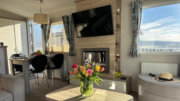 Uma sala de estar moderna com televisão de tela plana, lareira, área de refeições com cadeiras e vista para o mar.