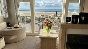 Uma sala de estar moderna com vista para a praia, com um banquinho redondo, uma mesa de centro de madeira com um vaso de flores, e uma área de cozinha com bancada preta e um fogão.