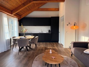 A modern kitchen with dark cabinets, a dining area with a round table and chairs, and a cozy living space with a sofa and a lamp. - Comfortable ground-floor accommodation for 2 persons with a private hot tub in the garden. Compact in size, yet warmly and fully furnished. (Otterlo)