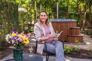 A person sitting at an outdoor table with a glass of wine, a vase of flowers, and a magazine. - Comfortable ground-floor accommodation for 2 persons with a private hot tub in the garden. Compact in size, yet warmly and fully furnished. (Otterlo)