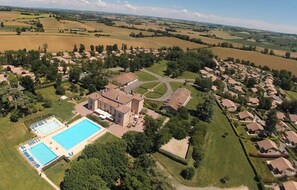 Une vue aérienne d’un quartier résidentiel avec une grande maison dotée d’une piscine, entourée de maisons plus petites et de champs ouverts.