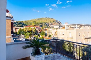 Ein Balkon mit einem Blumentopf, der auf eine Straße mit parkenden Autos und Gebäuden blickt, und ein Hügel mit Grün im Hintergrund.