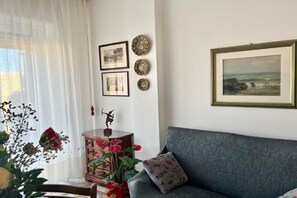 Un salon avec un canapé gris, une table d’appoint en bois surmontée d’une statue et un vase de fleurs. Des tableaux encadrés ornent le mur, et une fenêtre est agrémentée de rideaux blancs.