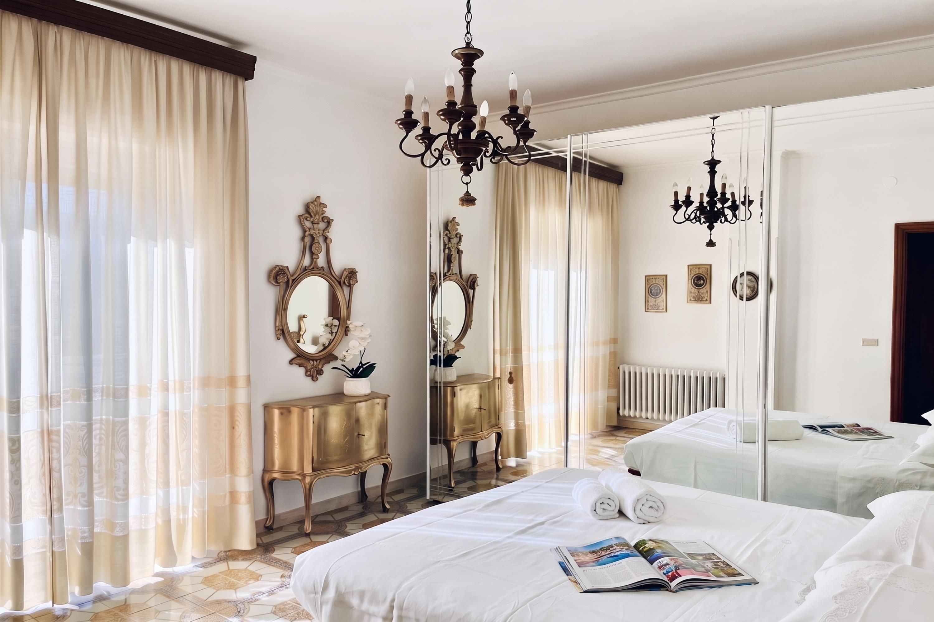 Une chambre d’hôtel avec un grand lit, deux tables de chevet, un miroir, un lustre et une vue sur l’extérieur par une fenêtre ornée de rideaux légers.