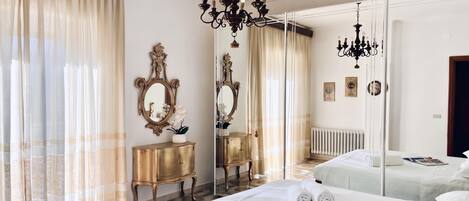 Une chambre d’hôtel avec un grand lit, deux tables de chevet, un miroir, un lustre et une vue sur l’extérieur par une fenêtre ornée de rideaux légers.
