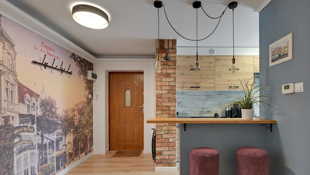 A hallway with a wooden floor, a wall mural, a wooden door, a brick accent wall, a kitchen area with a wooden countertop, two red stools, and pendant lighting. - Sopot 2 by Grand Apartments (Sopot)