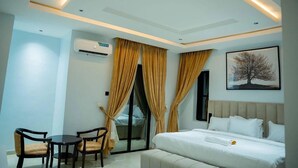 A modern hotel room with a bed, a desk with two chairs, a window with curtains, and a wall-mounted air conditioner. - Nirvana City Hotel (Benin City)